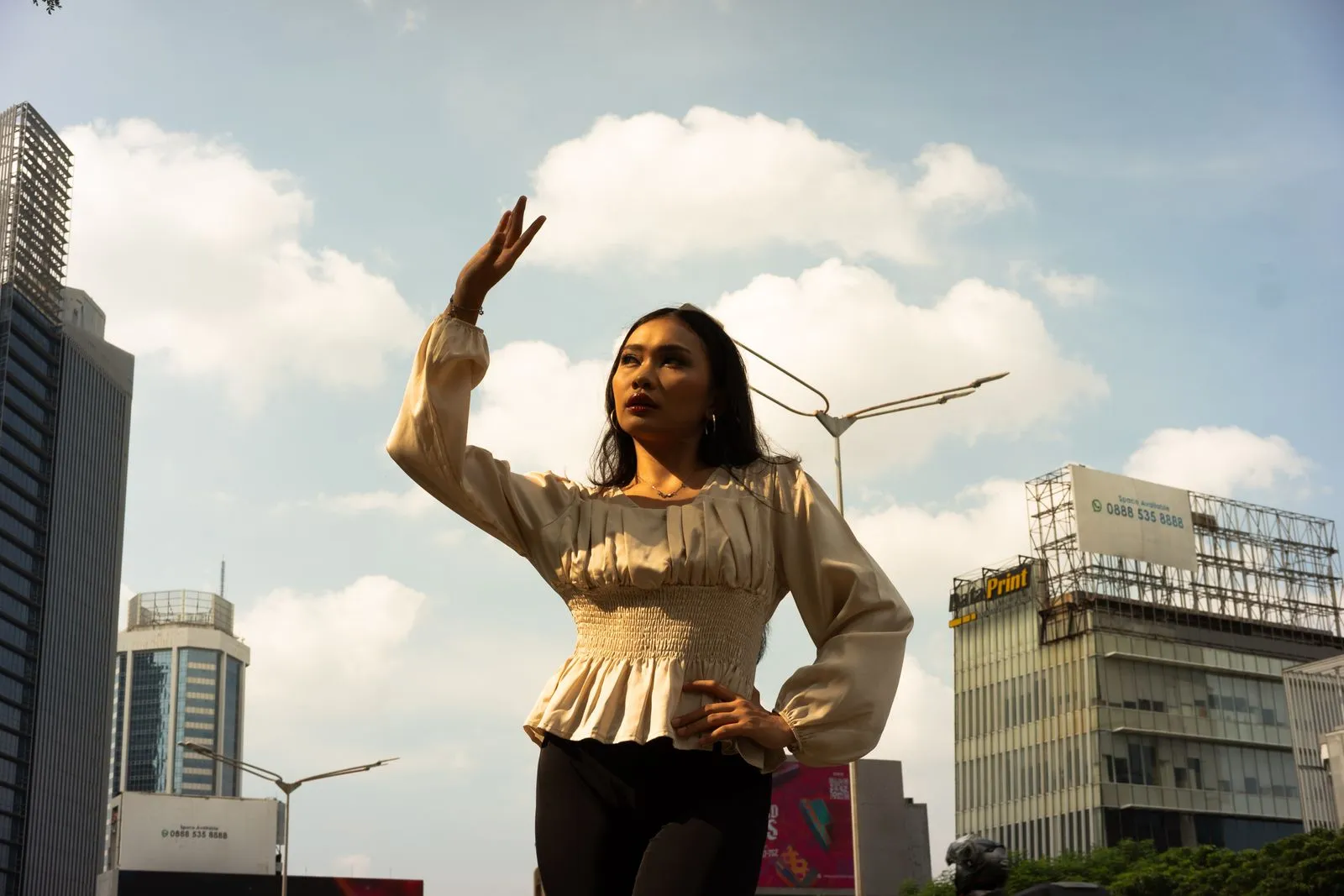 Woman raises hand confidently against city backdrop, showcasing modern urban fashion.